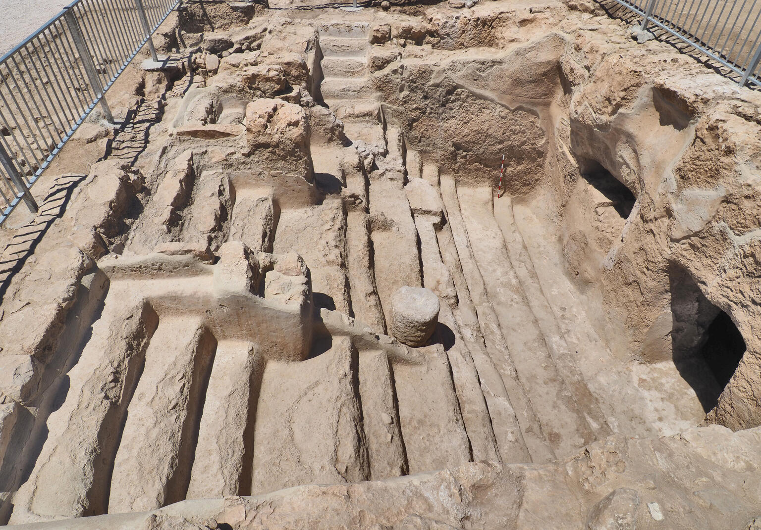 Ancient stone steps leading down into an area with vertical walls and some openings, surrounded by a metal fence. The area shows layered, weathered stone surfaces.