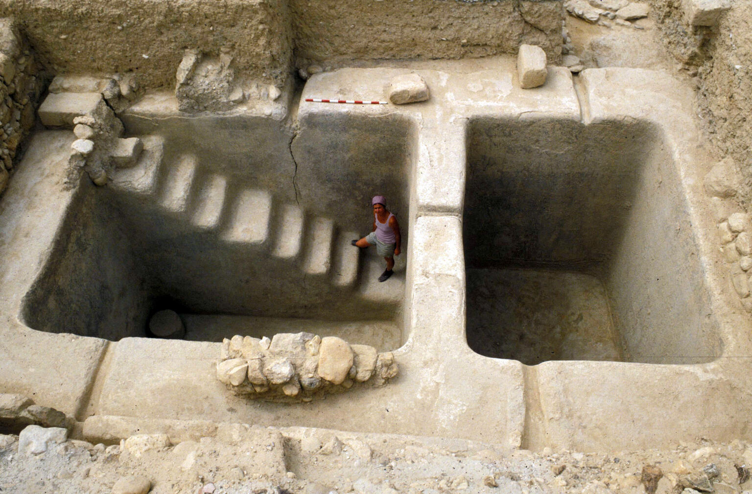 A person stands in a large, rectangular, stone excavation site with two deep pits, one with carved steps descending into it. The surrounding area is dry and sandy, with some scattered rocks.