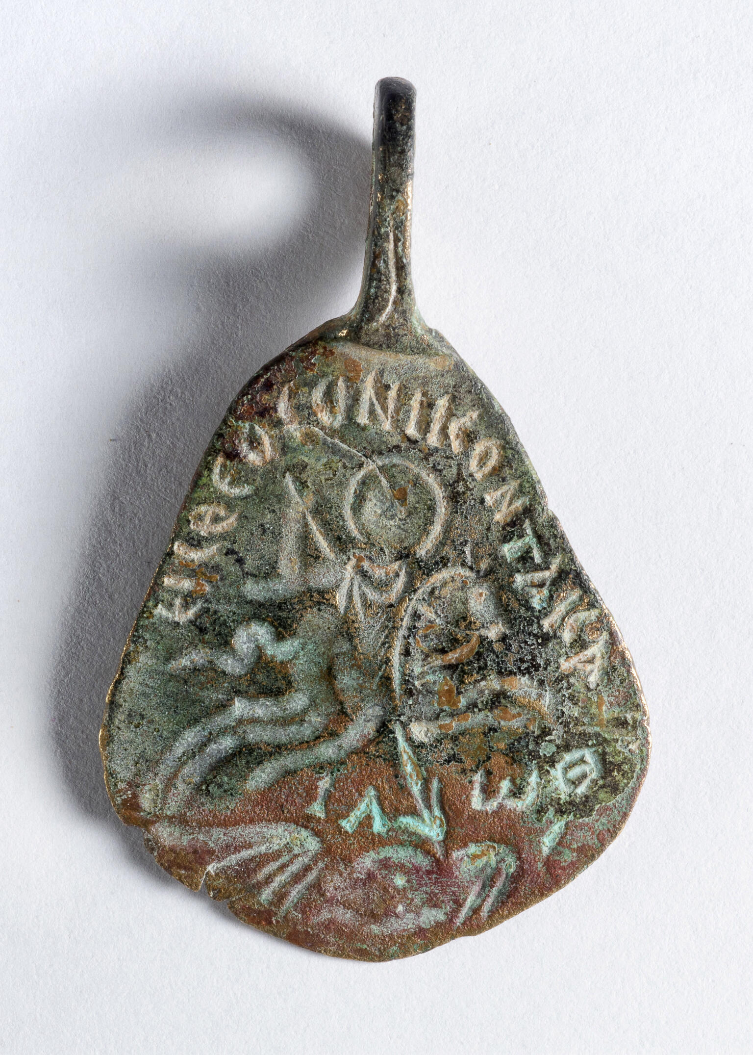 A weathered metal object with a triangular shape, featuring faint engraved text and an image of a horse and rider. The surface shows signs of age and patina, with a hook or loop at the top.