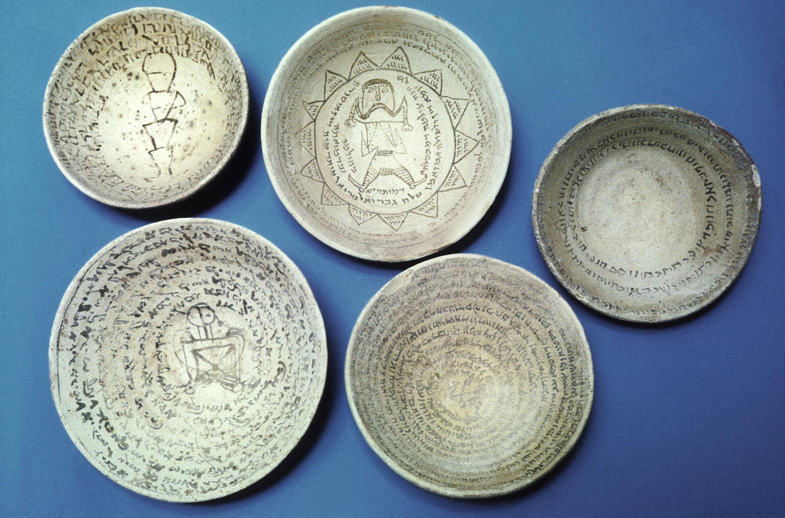 Five ancient ceramic bowls with inscriptions in a Semitic script, written in concentric circles. Some bowls feature geometric shapes and human-like figures.