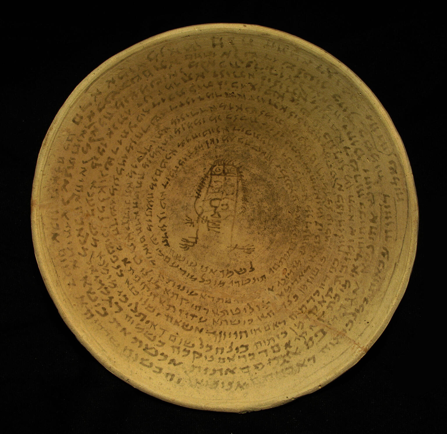 A shallow, round clay bowl with an inscription in a Semitic script written in concentric circles surrounding a central drawing of a human-like figure.