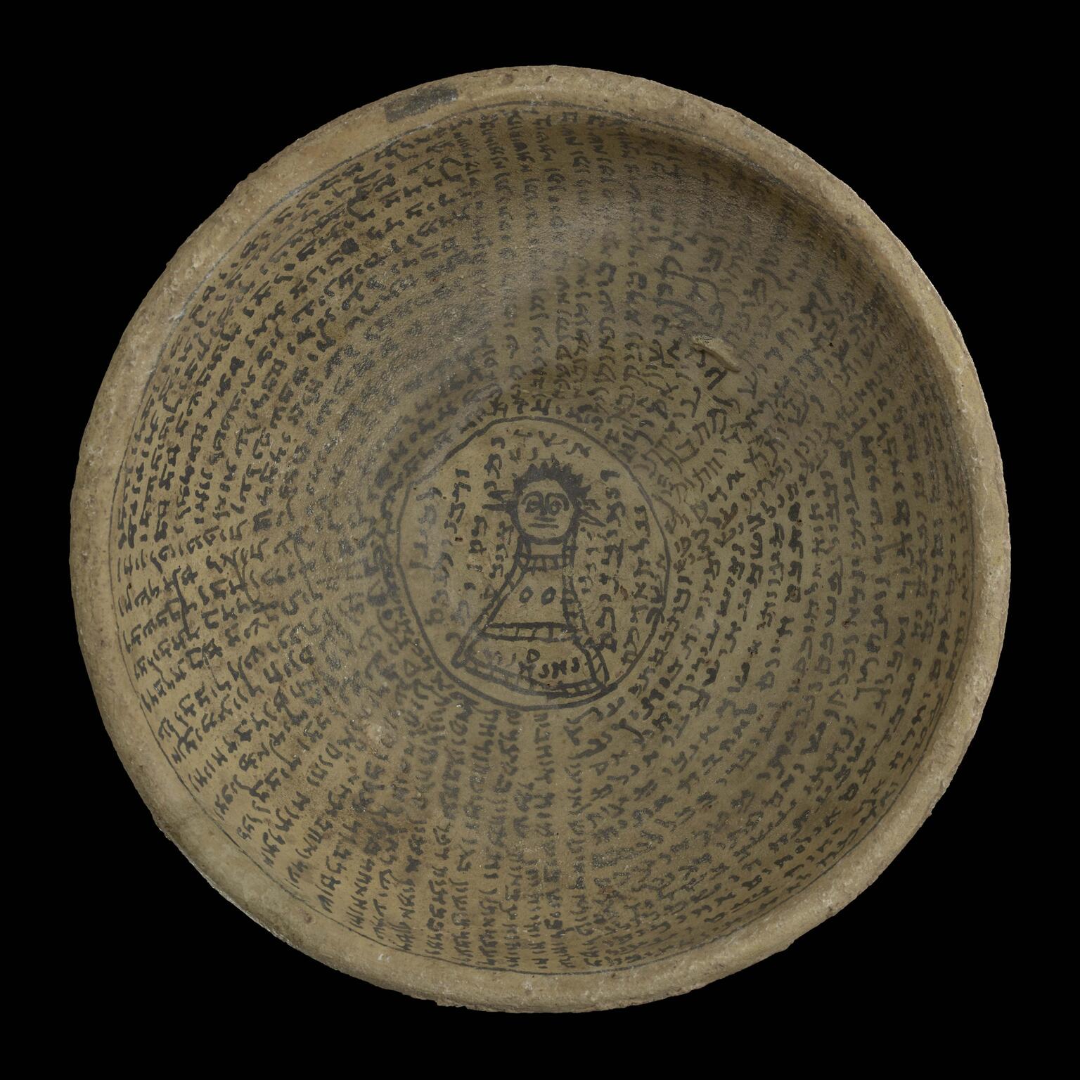 A round, ancient clay bowl inscribed with lines of text, some concentric and some radial, in a dark Semitic script. In the center is a simple drawing of a figure with wide eyes, surrounded by additional writing.