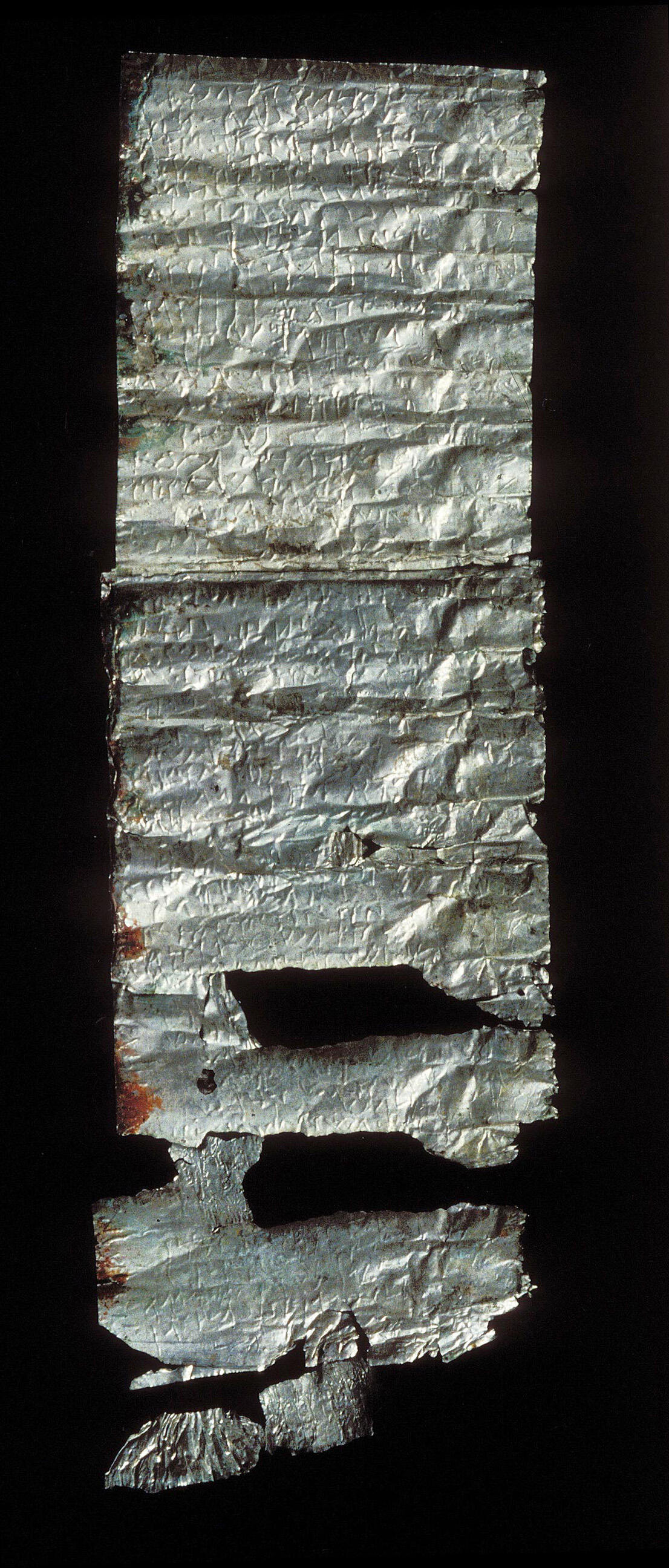 A fragment of an ancient, partially damaged silver scroll with faint inscriptions. Parts of the scroll are torn and missing.