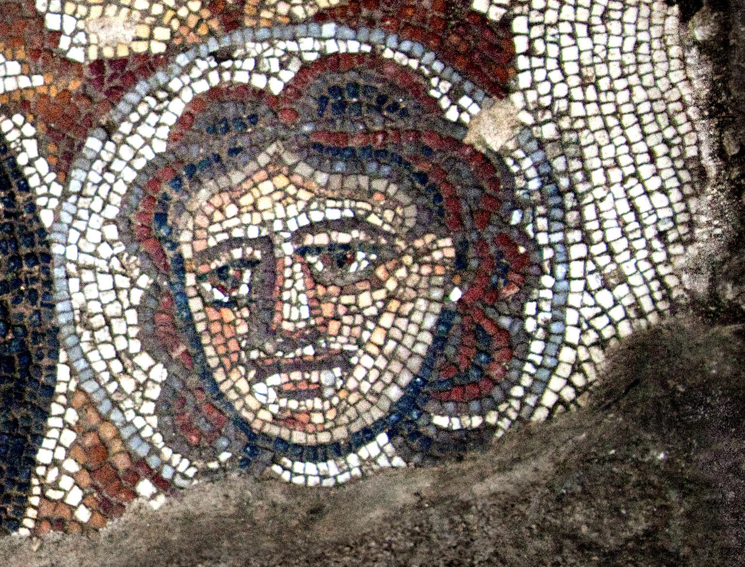 Ancient mosaic depicting a human face with red hair, large eyes, and a neutral expression, made from small colored tiles. The face is outlined with a halo-like border.