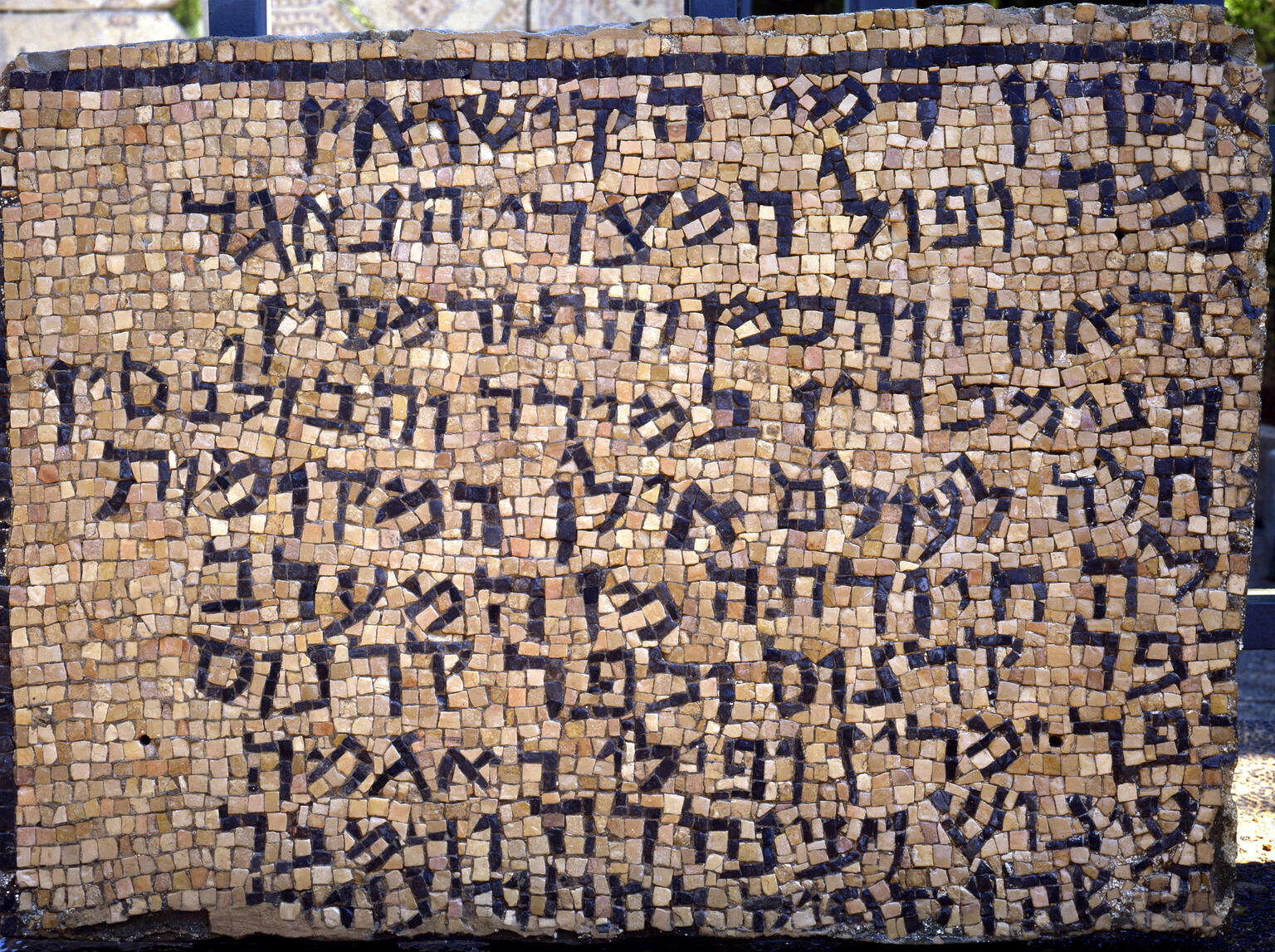 A rectangular stone mosaic with multiple lines of Hebrew script in dark tiles, set against a background of lighter-colored tiles. The mosaic appears weathered and is displayed outdoors.