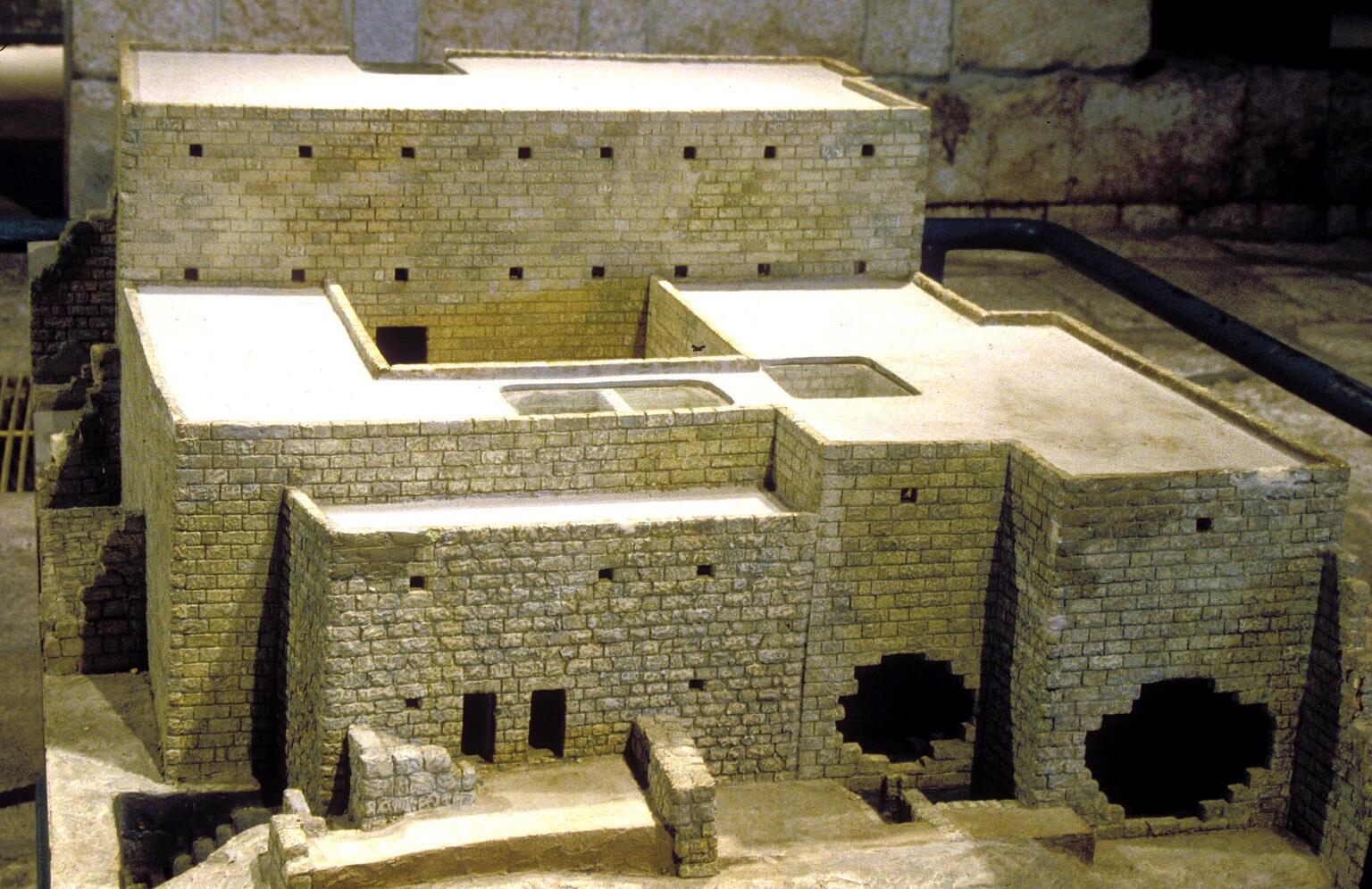 A detailed scale model of an ancient stone building featuring rectangular walls and small window openings, displayed against a stone backdrop.