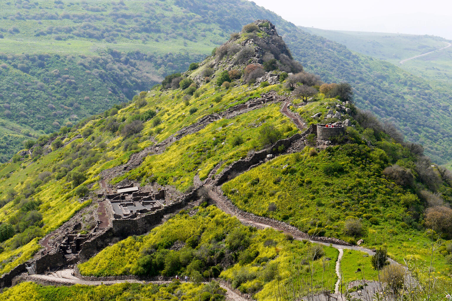Ruins of an ancient stone fortress with winding walls built along a lush, green hillside.