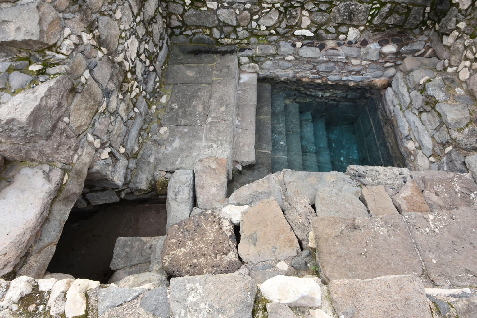 A stone structure with steps leading down into a rectangular pool of water, surrounded by weathered stone walls and blocks.