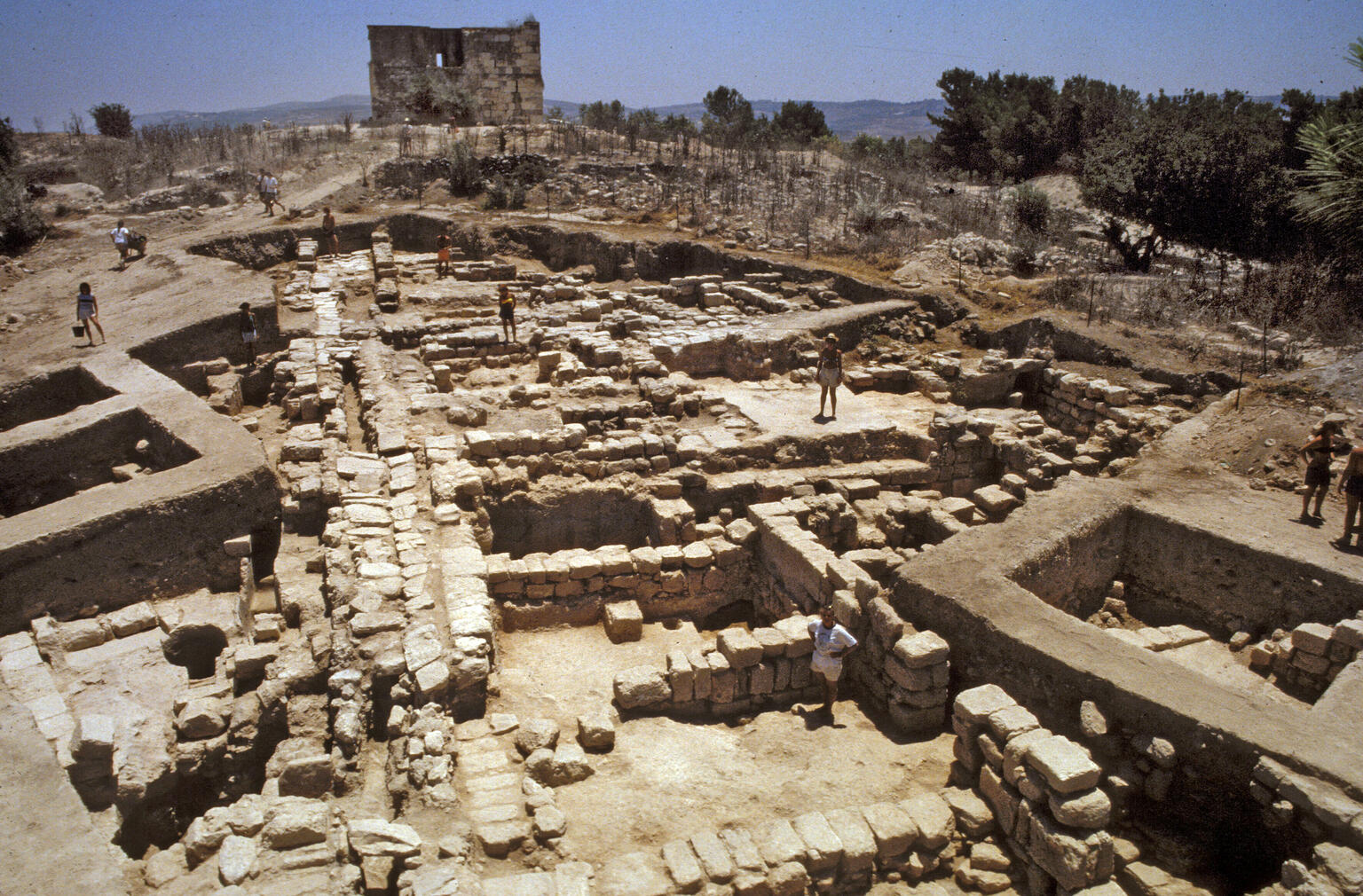 Ancient stone ruins with several people exploring the area; a partially intact building stands in the background under a clear blue sky.