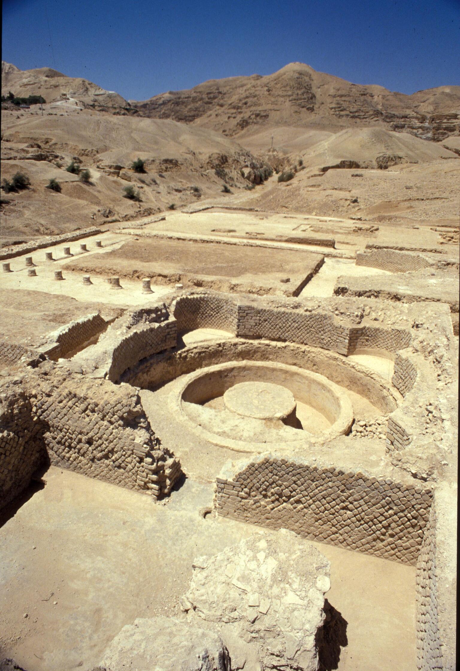 Ancient ruins with stone walls and a circular structure, set in a desert landscape with barren hills in the background under a clear blue sky.