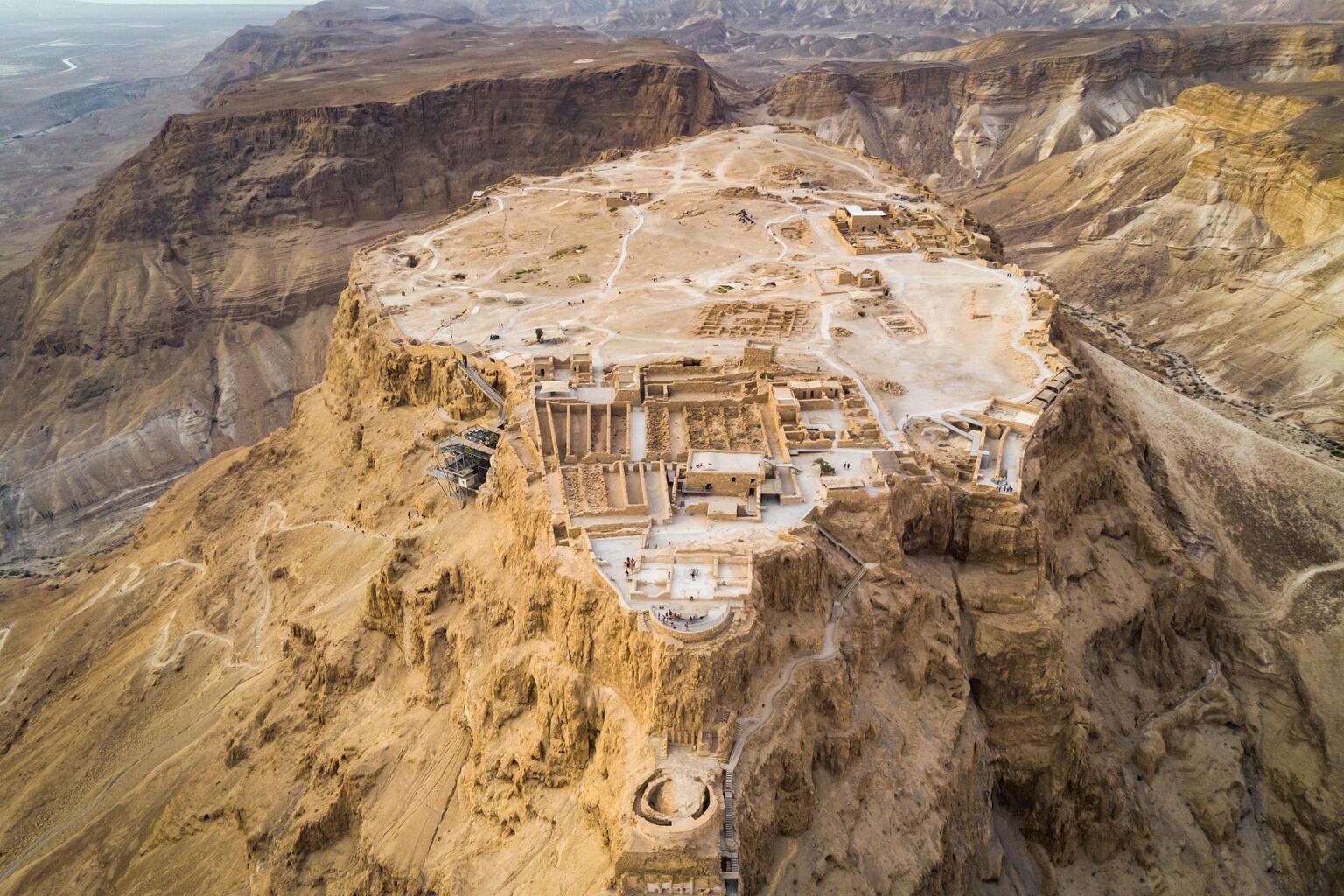 Aerial view of an ancient fortress, perched atop a rugged, rocky plateau in a desert landscape with scattered ruins and surrounding mountains.