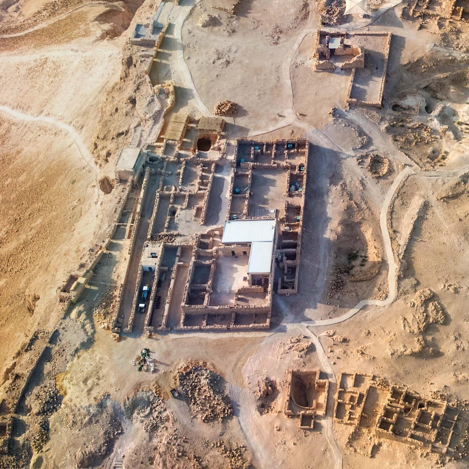 Aerial view of ancient ruins in a desert landscape, showing rectangular stone foundations, partially standing walls, and pathways winding around the site. The terrain is arid and rocky with sparse vegetation.