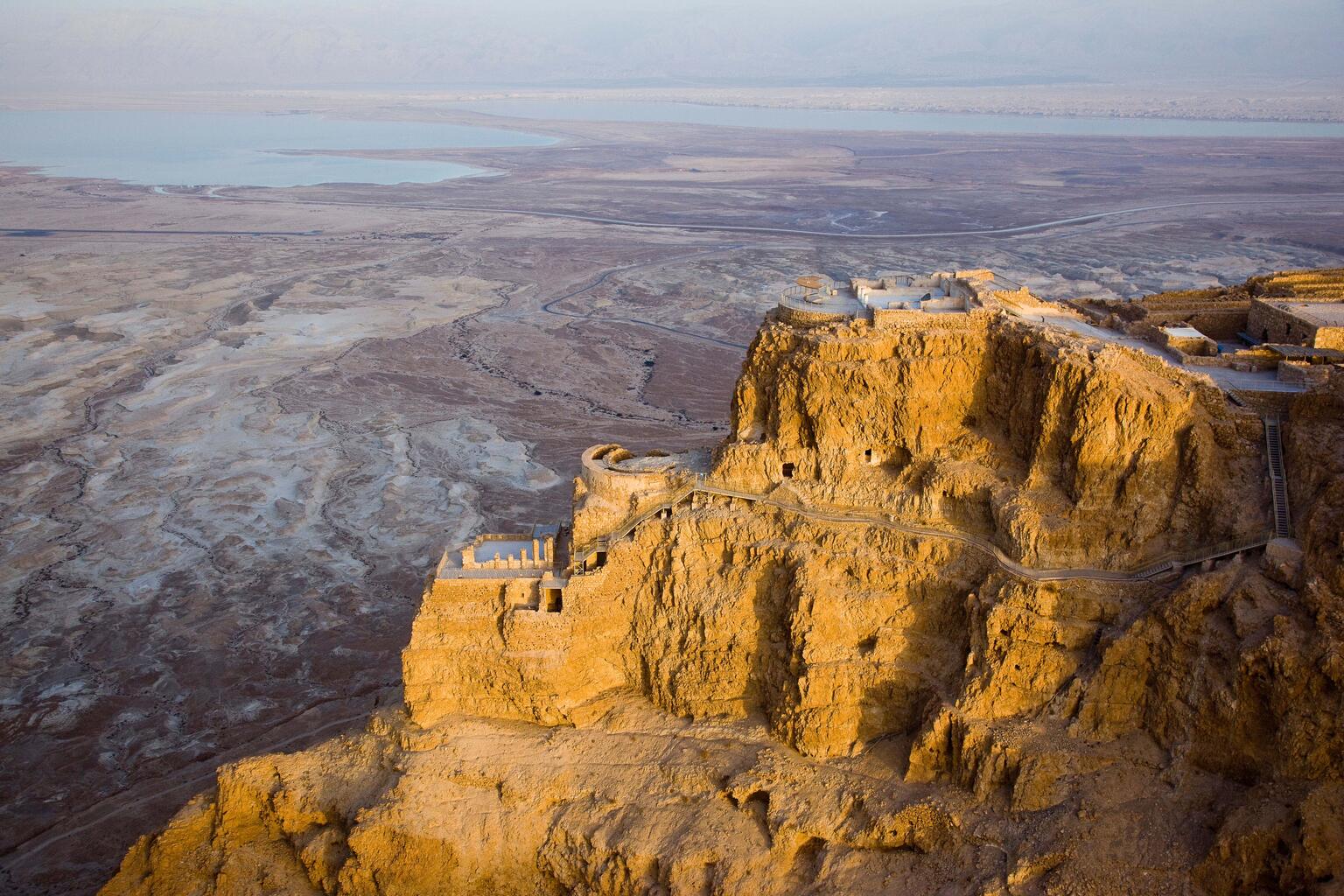 Aerial view of an ancient fortress atop a rugged desert plateau, with dramatic cliffs leading down to the dry landscape and the Dead Sea visible in the background.
