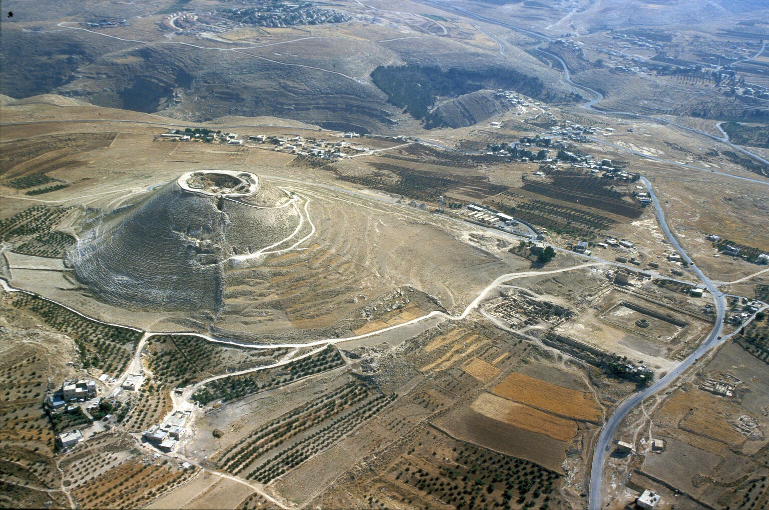 Aerial view of a rounded, steep-sided hill with ruins on top, surrounded by arid land, fields, scattered trees, roads, and small settlements in a hilly landscape.