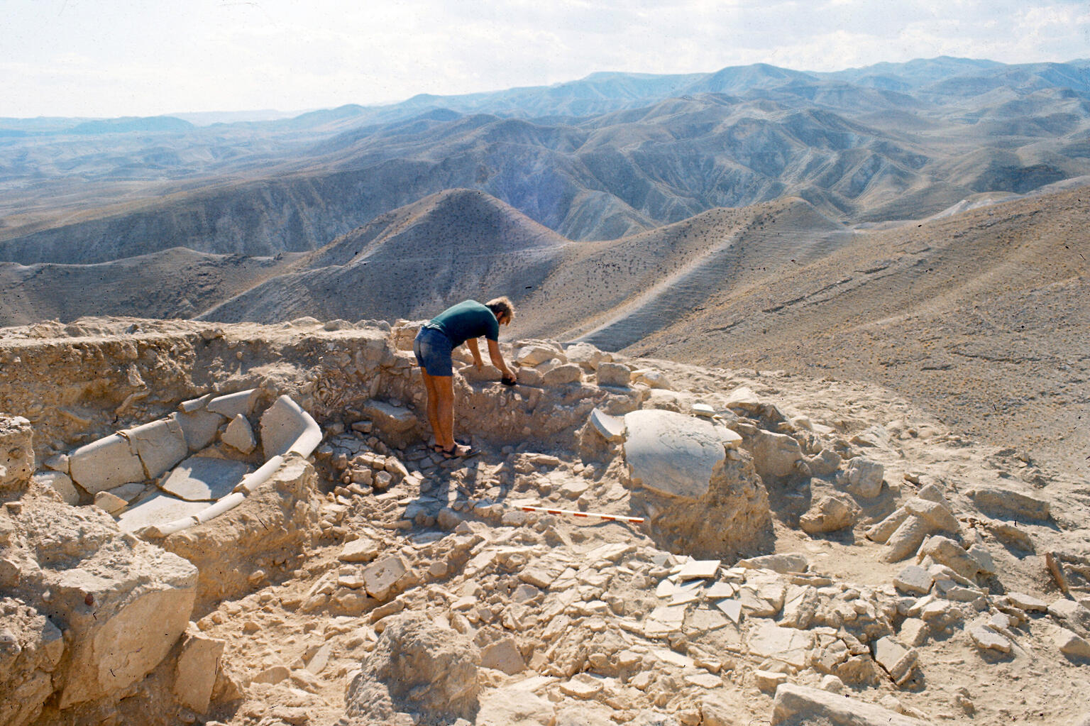 A person examines ancient stone ruins atop a rocky hill, surrounded by a vast, arid mountain landscape under a partly cloudy sky. Archaeological tools are scattered nearby.