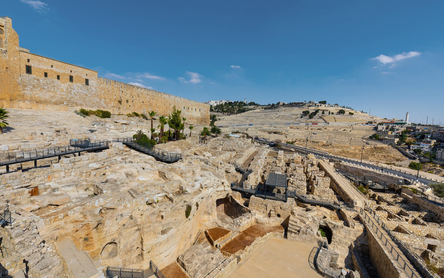 Ancient stone ruins and archaeological site near a large stone wall, with hills and scattered buildings in the background under a clear blue sky.
