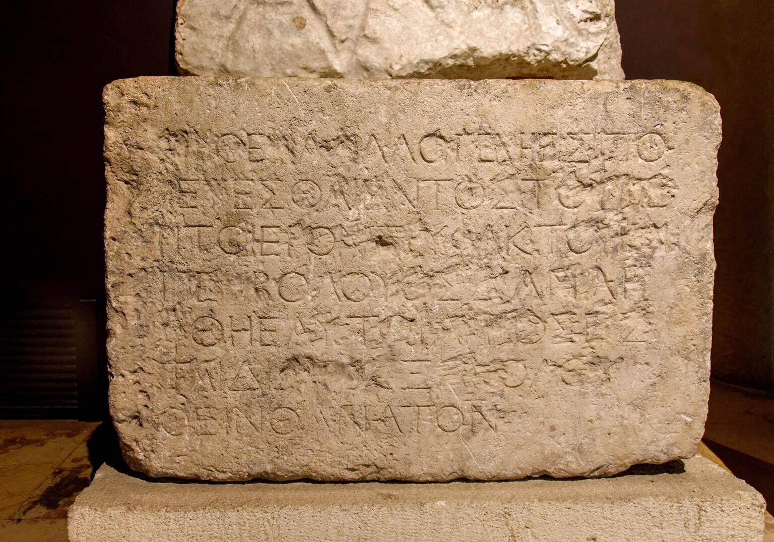 A rectangular ancient stone tablet with worn Greek inscriptions carved into its surface, displayed upright in a museum setting. The text is faded and partially eroded, making some letters difficult to read.
