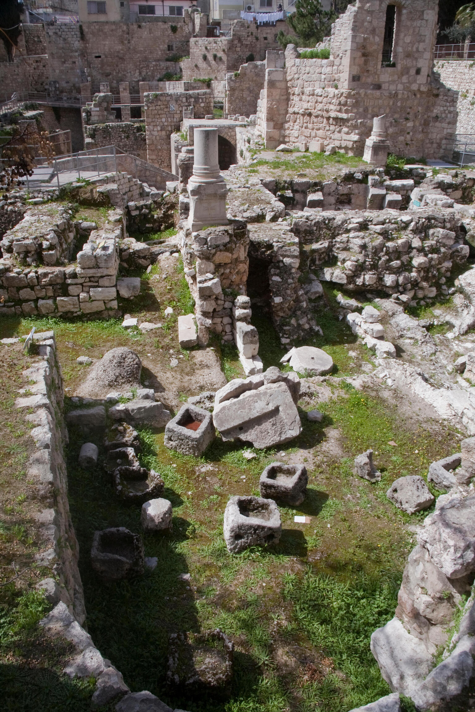 Ancient stone ruins with broken columns and scattered stone blocks, surrounded by moss and grass, set within ancient stone walls and partially preserved structures.