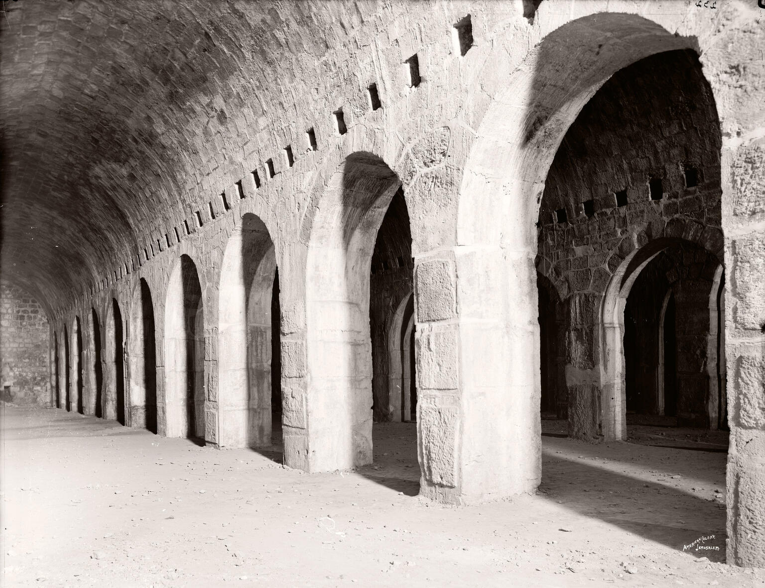 A black-and-white photo of a stone building interior shows a row of large, evenly spaced arched doorways under a vaulted ceiling, with light casting shadows on the sandy floor.