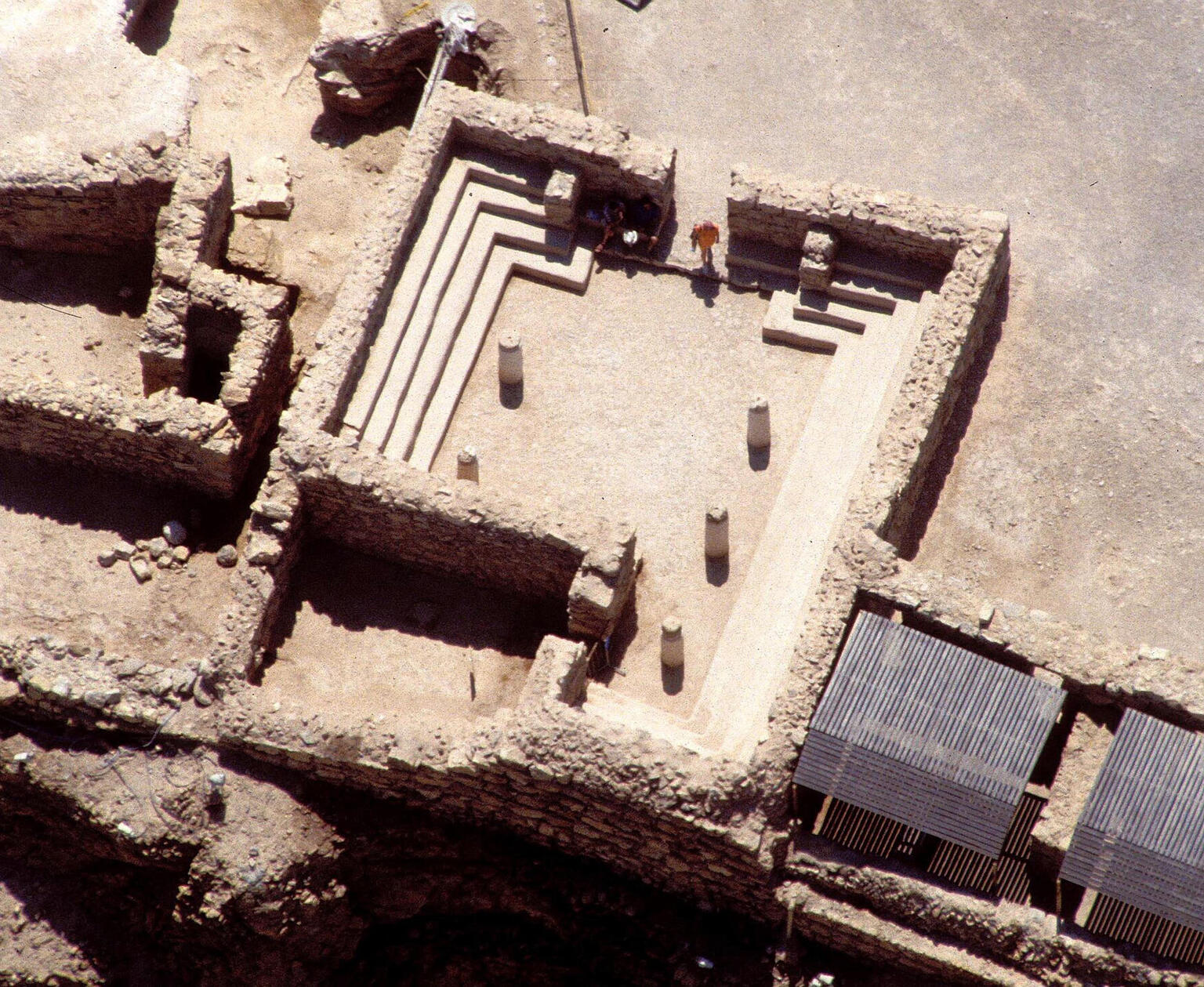 Aerial view of an ancient stone structure with steps along the sides, five central pillars, and partially intact surrounding walls in a desert archaeological site.