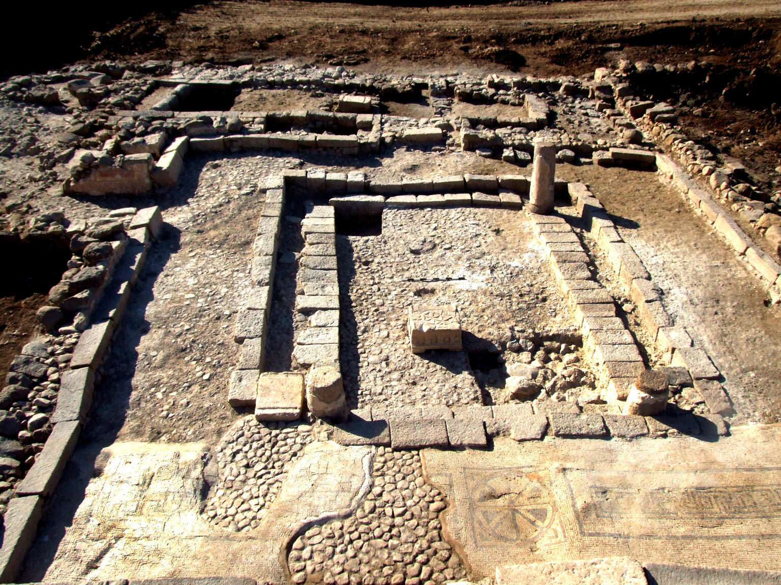 Aerial view of ancient stone ruins featuring rectangular walls, columns, and a mosaic floor with geometric patterns and symbols. The site is partially excavated, revealing the layout and textures of the structure.