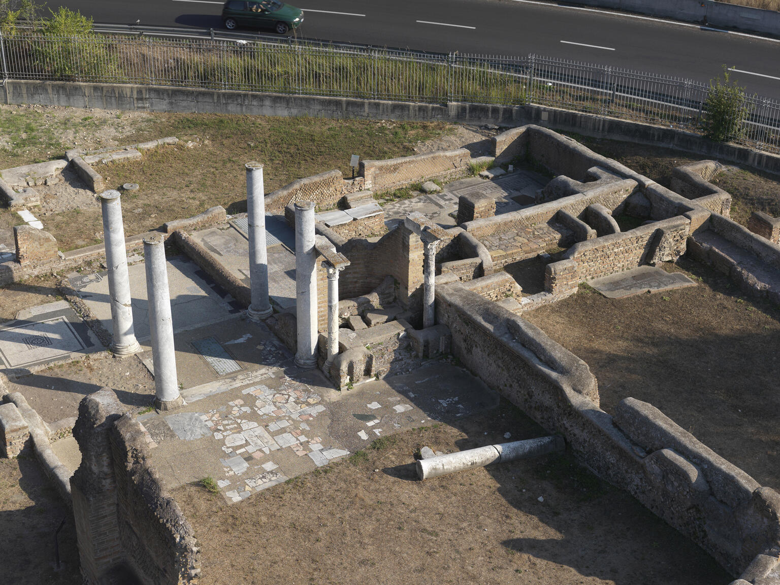 Aerial view of ancient stone ruins with several standing columns, broken walls, and scattered stone slabs, bordered by a fence with a road and car visible in the background.