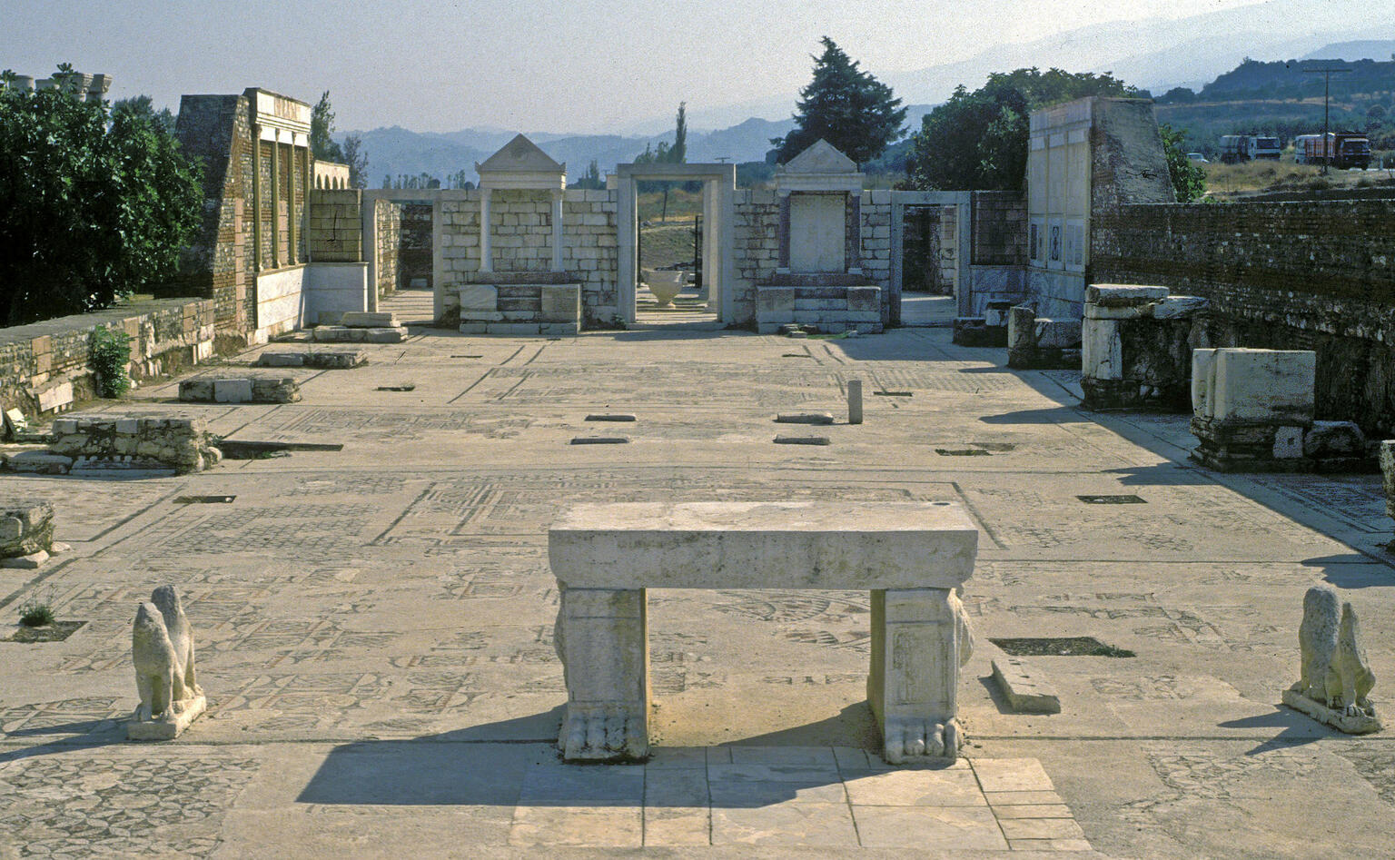 Ancient stone ruins of a large hall with mosaic floors and carved columns, open to the sky, surrounded by trees and hills in the background. At the near end of the hall is a two-legged table flanked by the remains of animal statues. At the far end is a doorway between two niches with triangular tops.