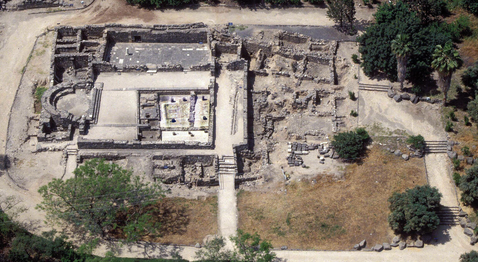 Aerial view of ancient stone ruins with rectangular and circular structures, surrounded by trees and pathways.
