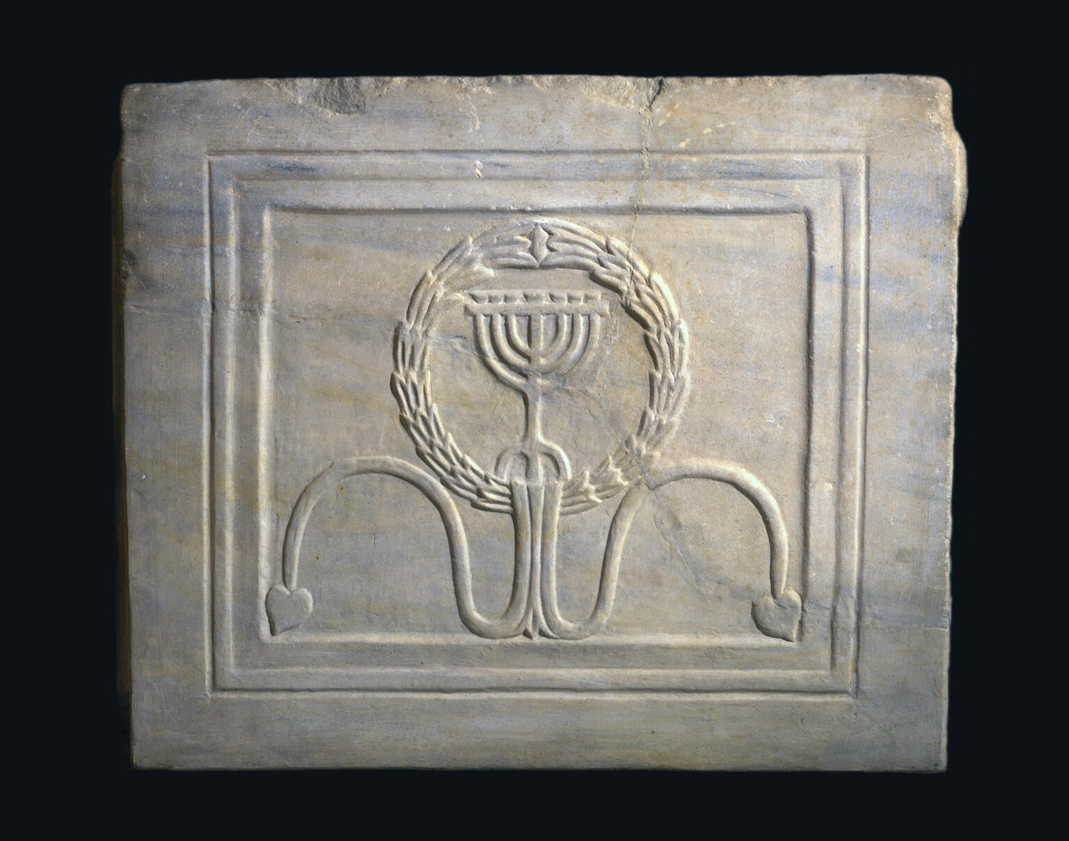 A carved marble tablet featuring a menorah inside a wreath, with two curved lines ending in leaf shapes beneath it. The design is framed by a rectangular border.