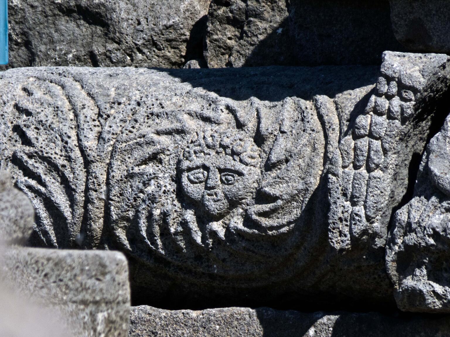 Close-up of an ancient stone carving featuring a face surrounded by radiating lines, with a partially preserved wing carved to the right of the face, set against a rough stone background.