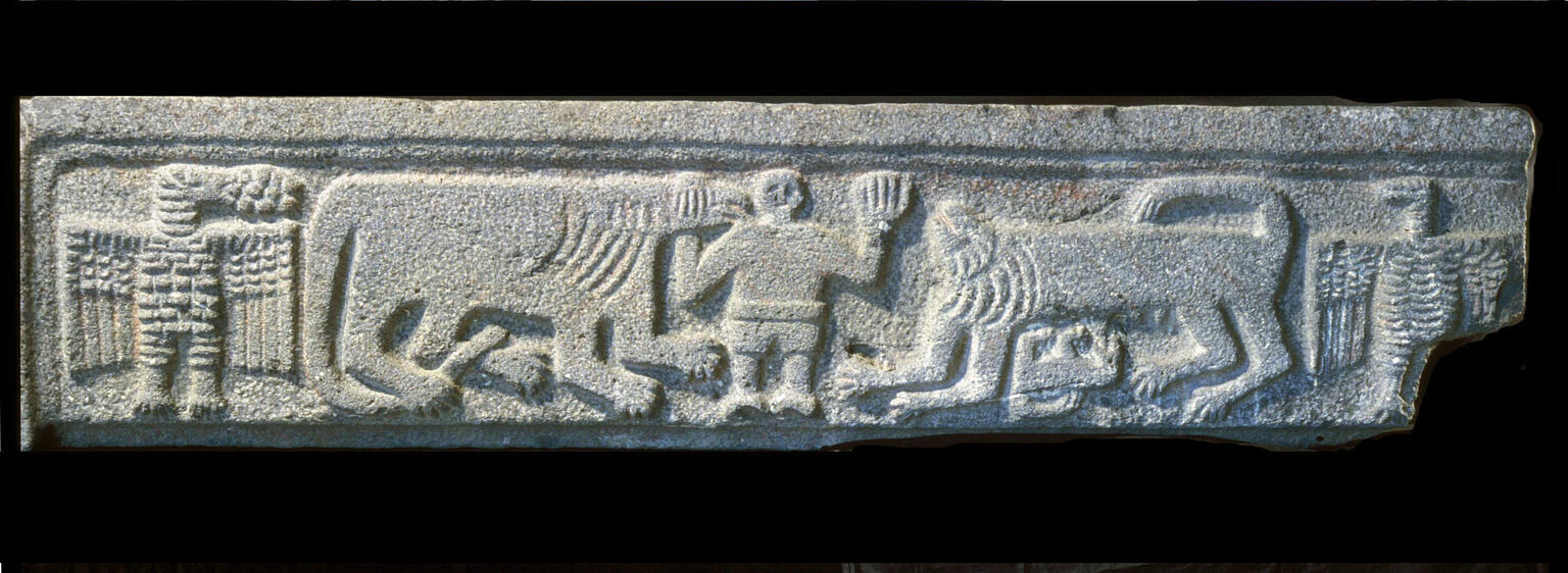 Ancient stone relief showing a human between two lions flanked by two eagles. A lion cub lying on its back nurses from the lion on the right.