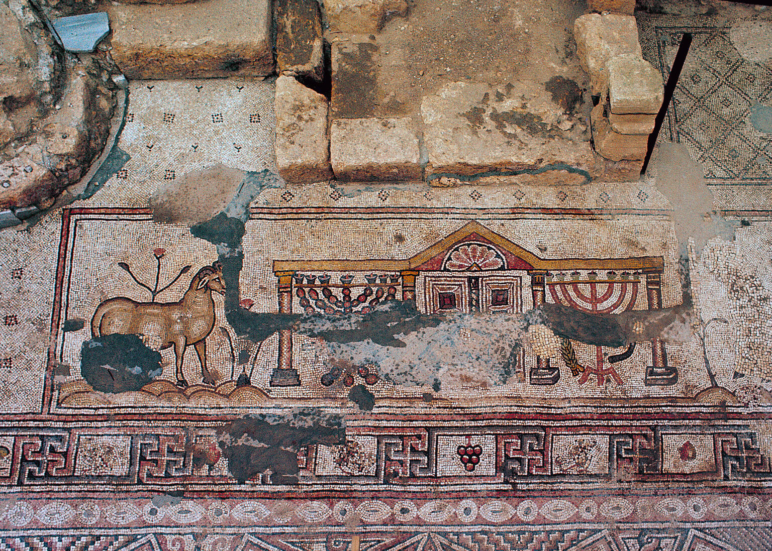 Ancient mosaic with a deer, a building with columns, menorahs, ritual objects, and geometric patterns, partially damaged and surrounded by stone ruins.