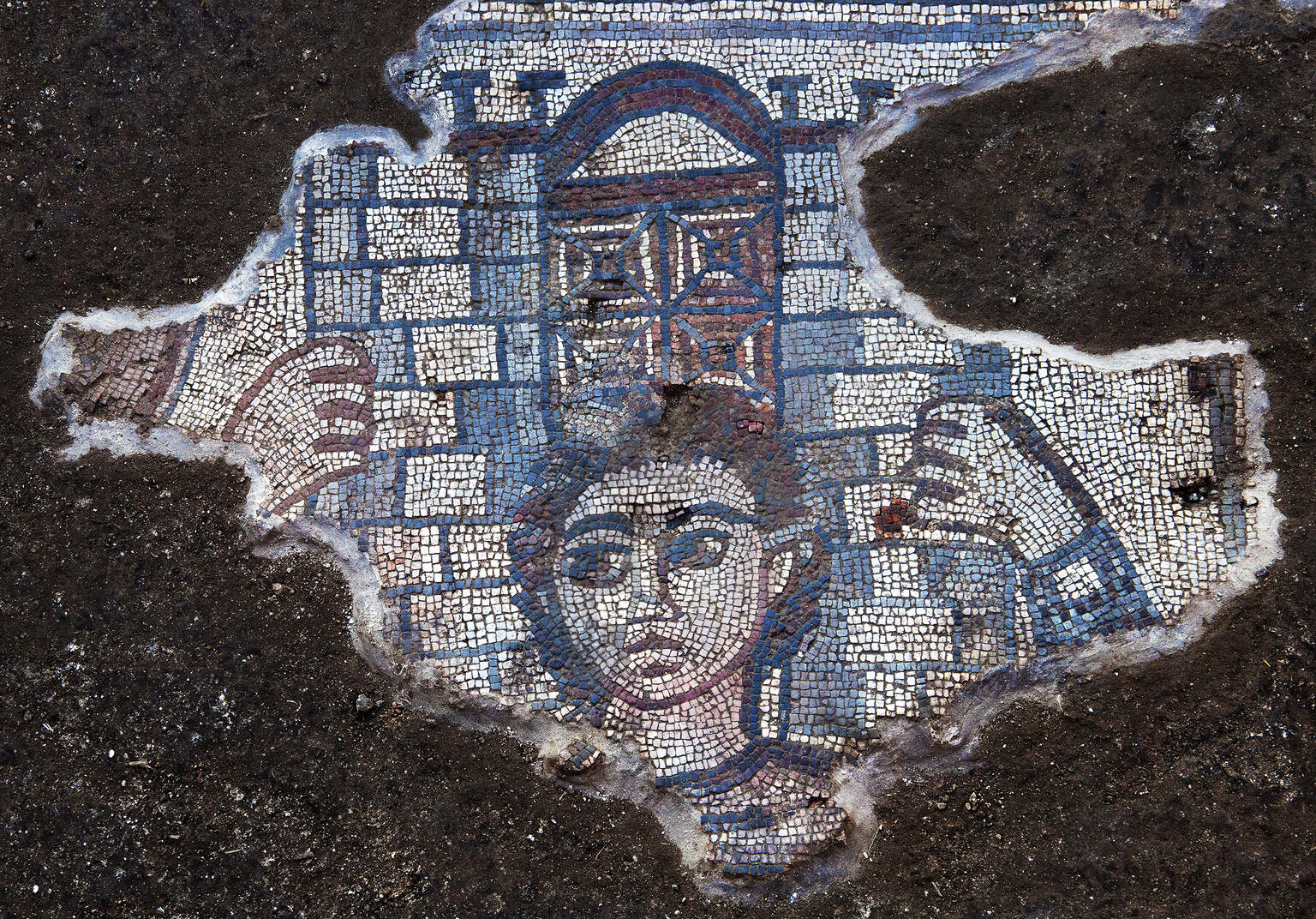 A fragment of an ancient mosaic depicts a human face and hands grasping an architectural structure with a gate in the center. The surrounding area shows dark, rough ground.