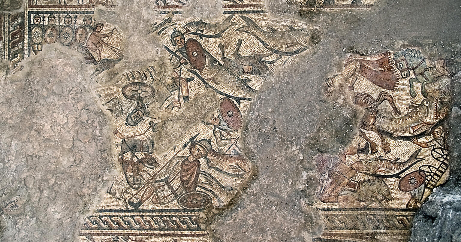 Ancient mosaic depicting armored figures, horses, and combat scenes; some parts of the artwork are missing or damaged, revealing patches of bare stone.
