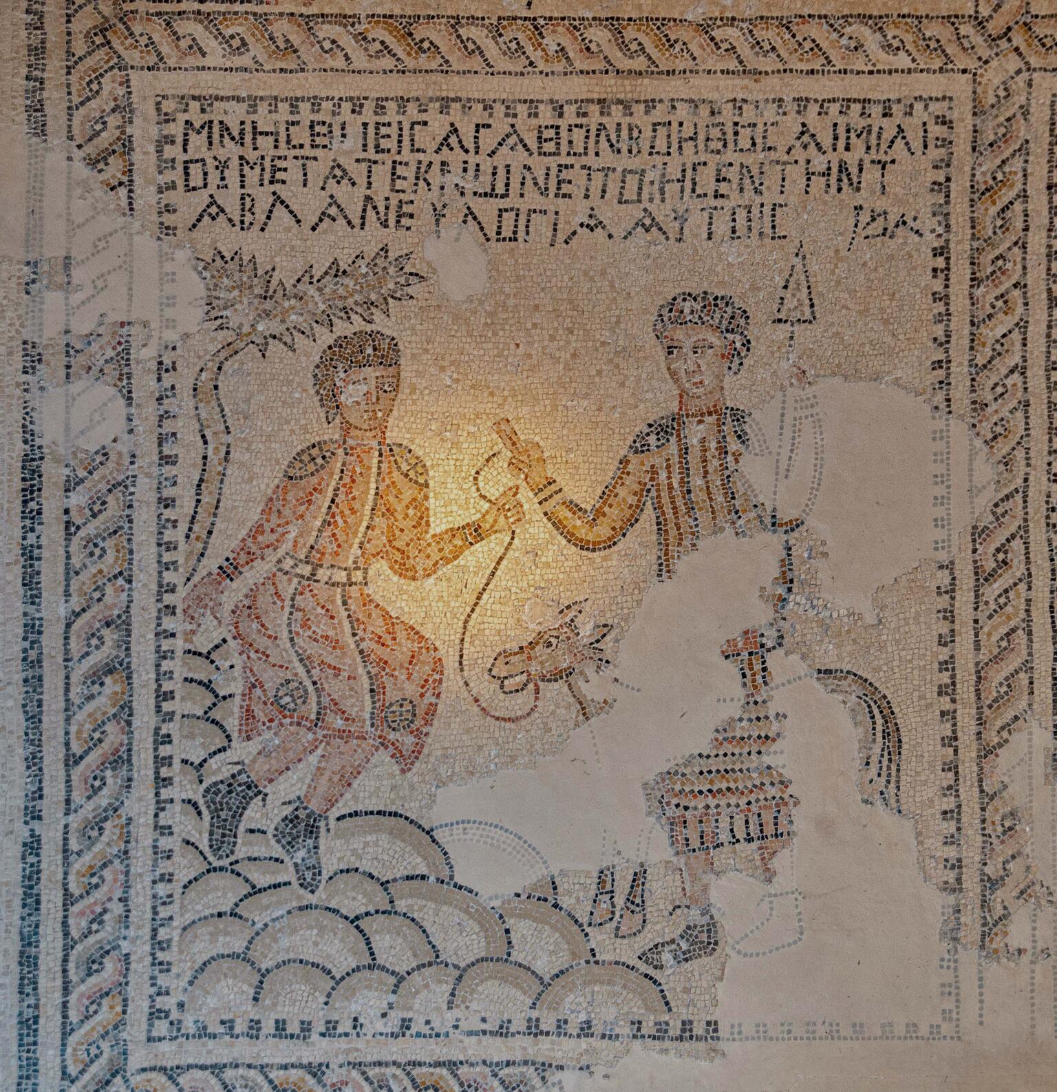 Ancient mosaic depicts two robed figures and a donkey. Greek text appears above the scene. There are areas of visible wear and missing tiles.