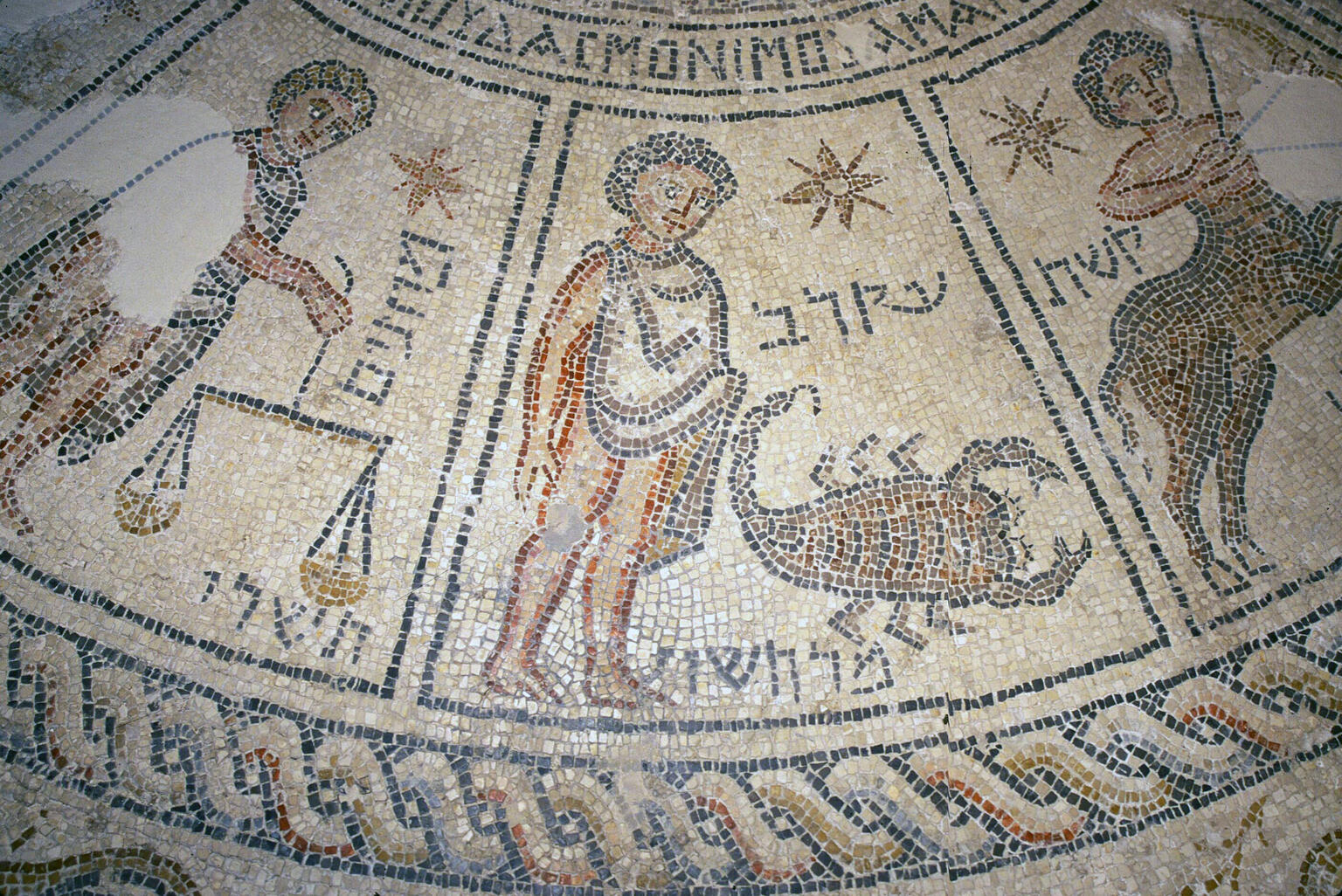 Ancient mosaic depicting three zodiac signs: Libra with scales, Scorpio with a scorpion, and Sagittarius as a centaur archer. Hebrew inscriptions and decorative patterns frame the figures.