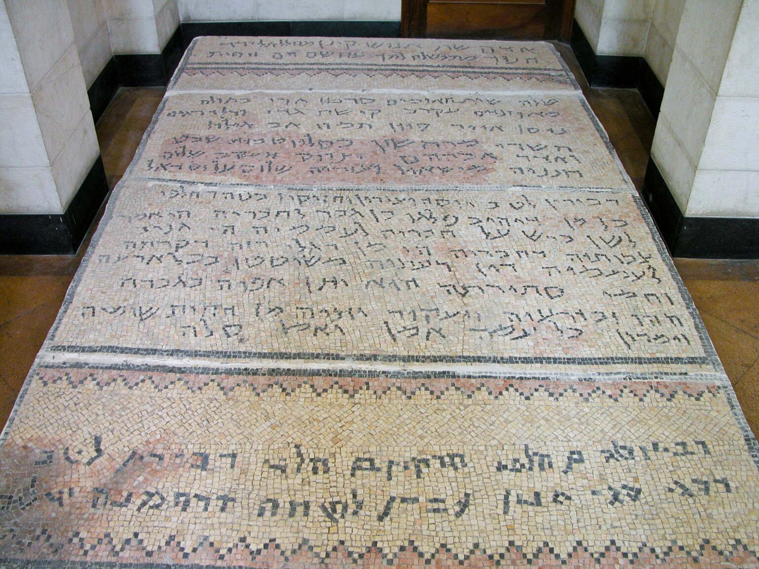A rectangular floor mosaic with Hebrew and Aramaic inscriptions in black letters, bordered by a geometric pattern, set in a tiled hallway with stone walls.