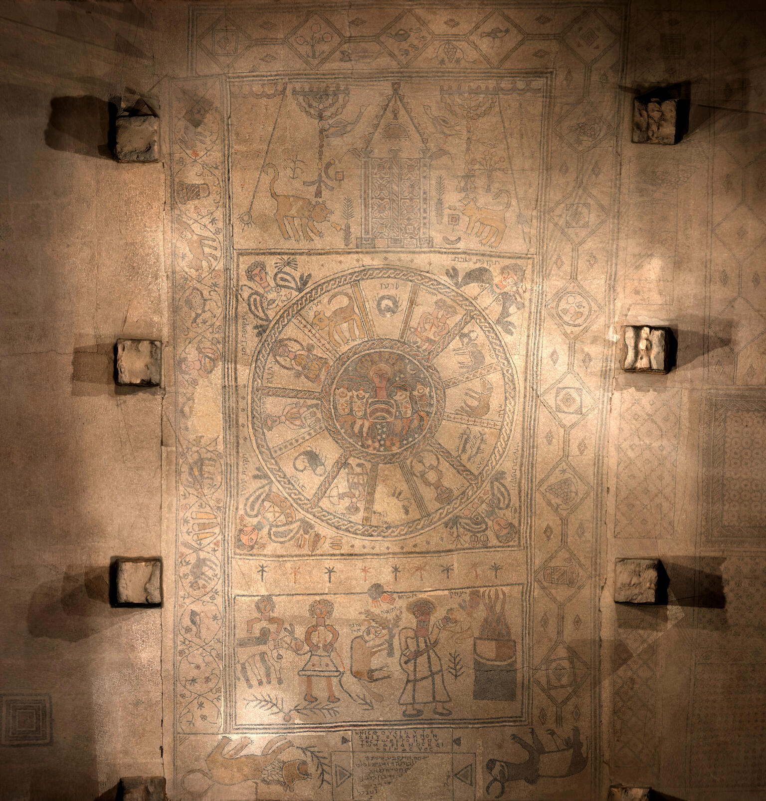 Mosaic panel with zodaic in the center, figures on the bottom, and ritual objects and animals on the top, surrounded by stone and several square blocks.