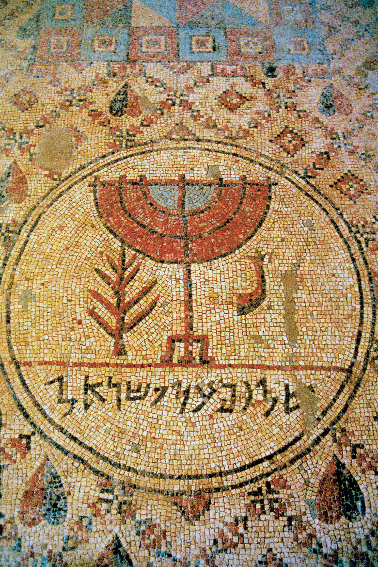 Ancient mosaic featuring a central menorah design, flanked by a plant and a shofar, with Hebrew text below, all set within a circular border amid intricate geometric patterns.