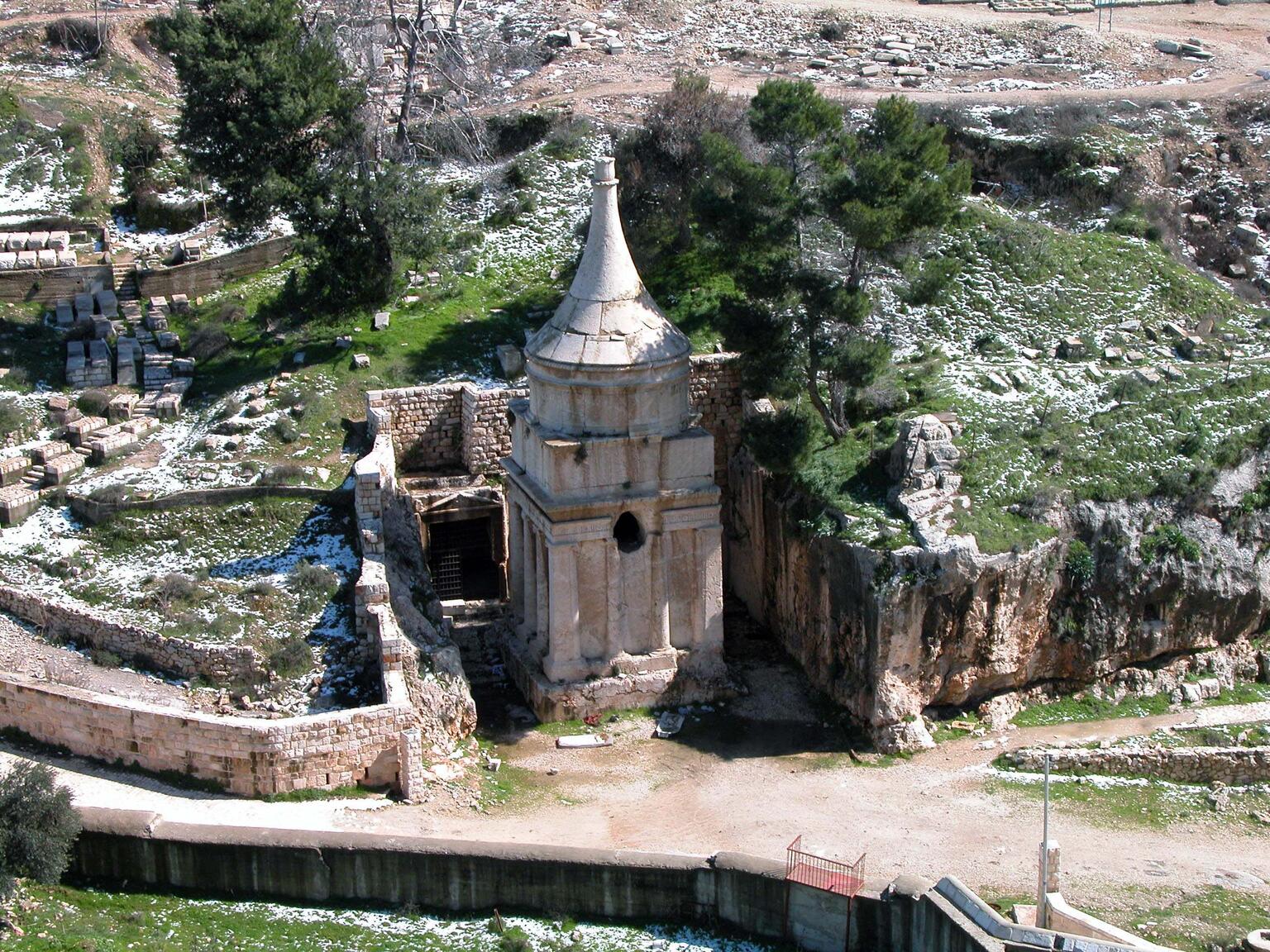 A stone mausoleum with a conical roof stands surrounded by rocky terrain, snow, and scattered trees, with ancient ruins visible nearby.