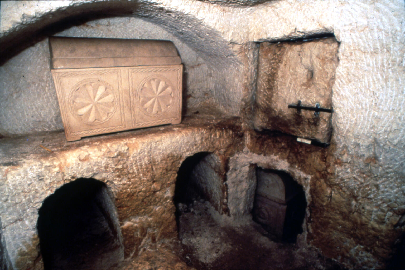 Ancient stone tomb with arched niches and a decorated limestone ossuary carved with rosettes, set in a small, dimly lit rock-cut chamber.