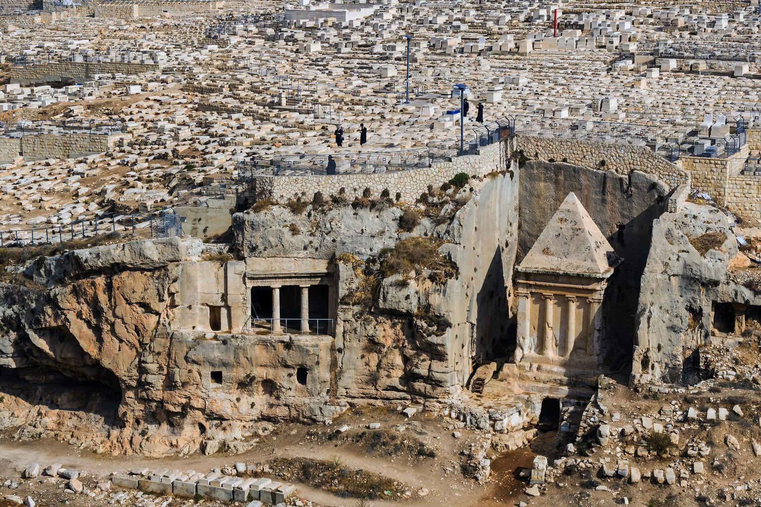 An ancient rock-cut tomb complex with rectangular openings and a pyramid-roofed structure stands in a vast hillside cemetery with countless stone graves and a few people.