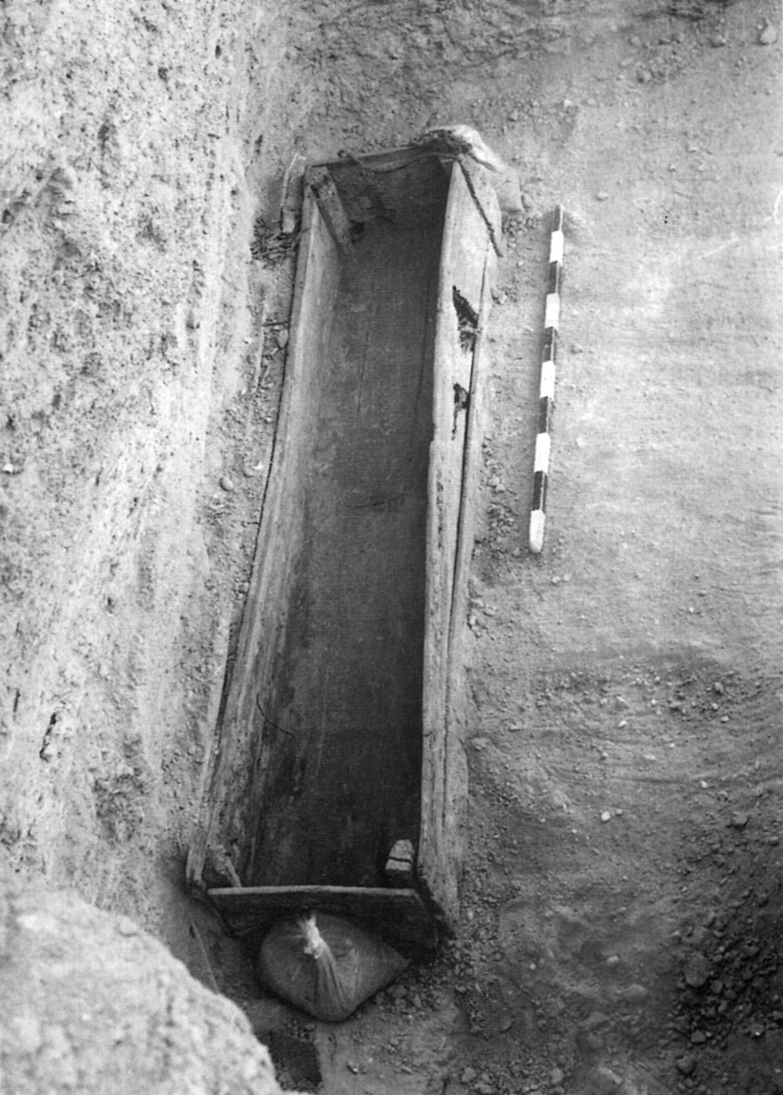 Black-and-white photo of a wooden coffin in an excavated grave site, seen from above, with a measuring stick placed beside it for scale and a sandbag at one end.
