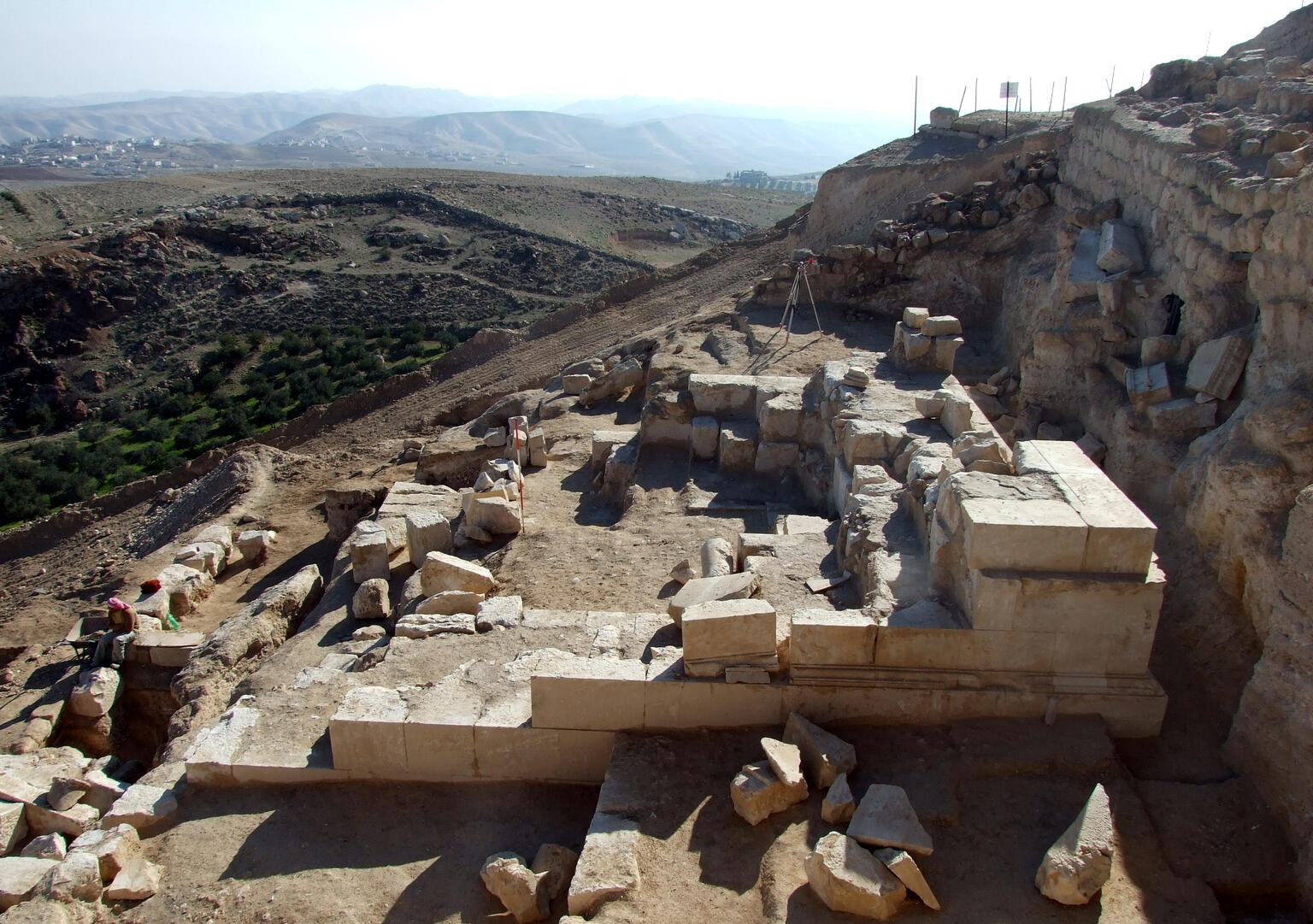 Ancient stone ruins at an archaeological site on a hillside, with scattered blocks and partial walls. Rolling hills and a distant settlement are visible in the background under a hazy sky.