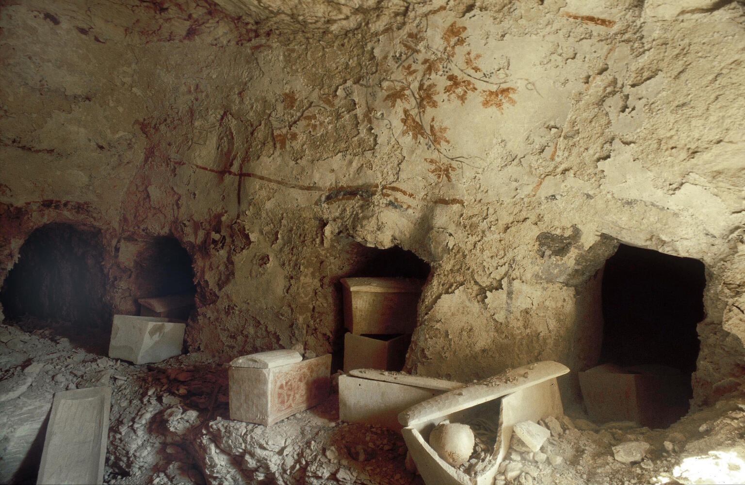 Ancient rock-cut tomb chamber with three burial niches in a rough limestone wall, several broken stone sarcophagi, and scattered debris on the ground. Faint painted decorations are visible on the wall.