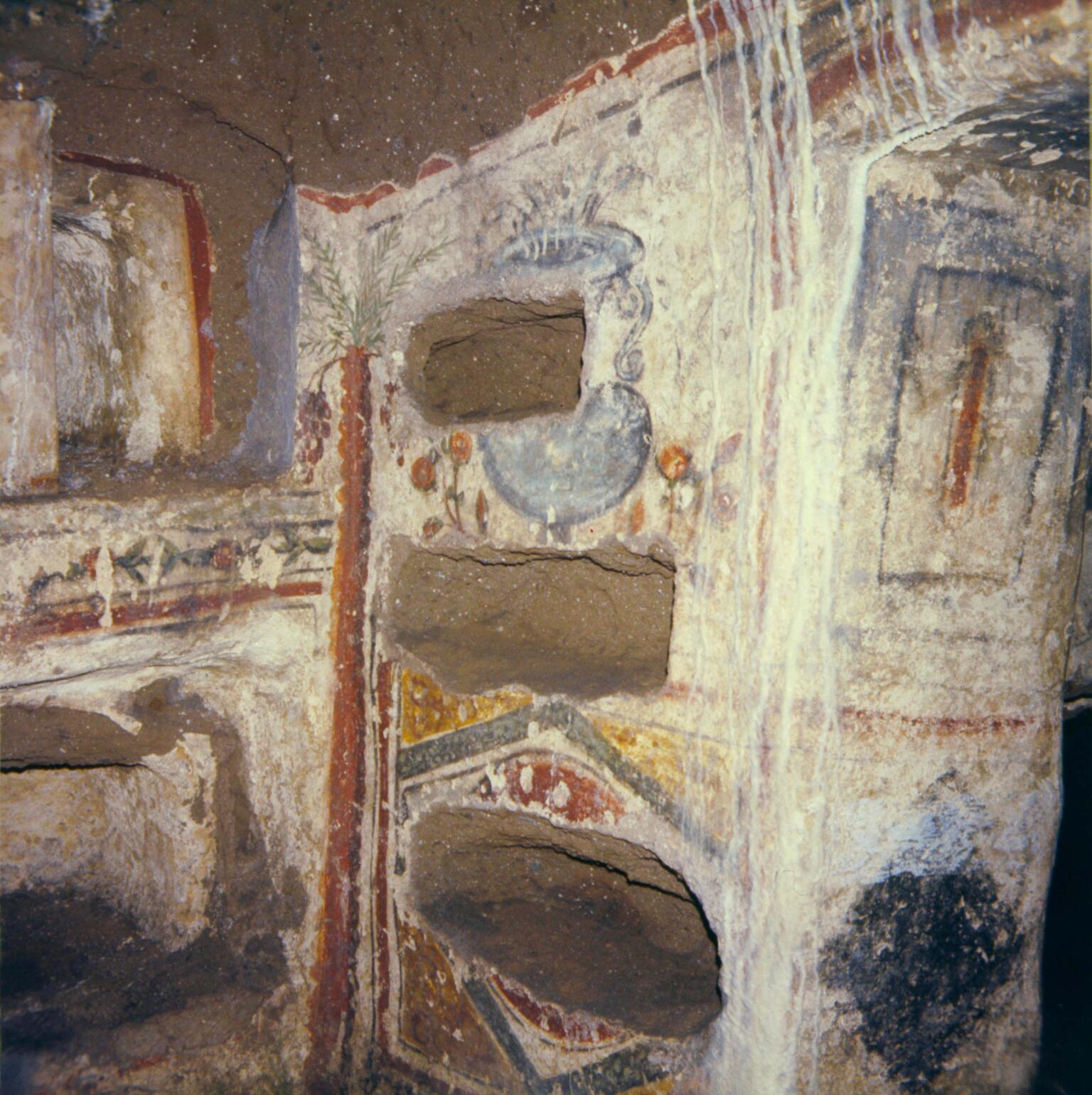 Ancient stone tomb with rectangular niches carved into the walls, decorated with faded frescoes and geometric patterns in red, yellow, and blue. Some niches appear empty; white mineral streaks run down the wall.