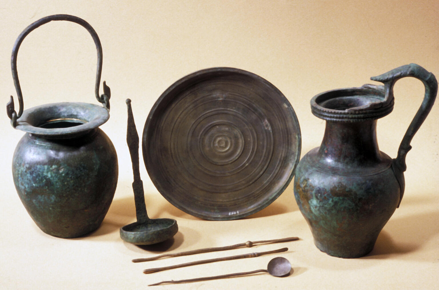 A collection of ancient bronze vessels and utensils, including two jugs with handles, a round plate, a ladle, and three long utensils.