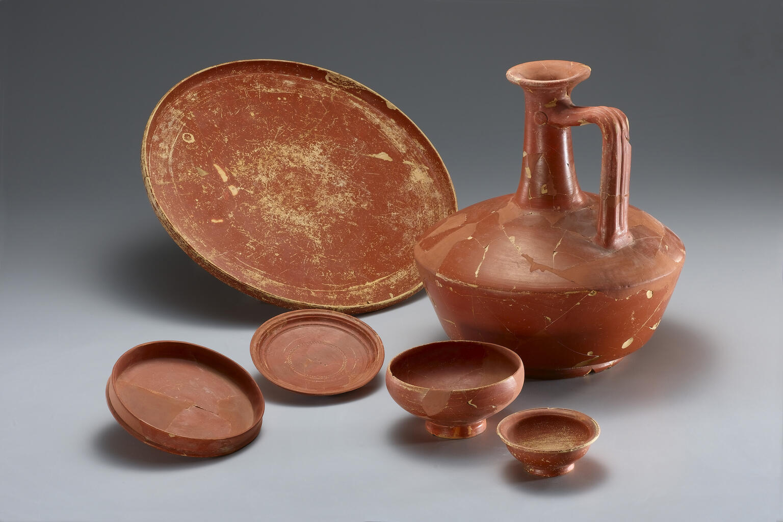 A collection of ancient red-glazed pottery, including a large jug with a tall neck and handle, a wide shallow dish, and four smaller dishes.