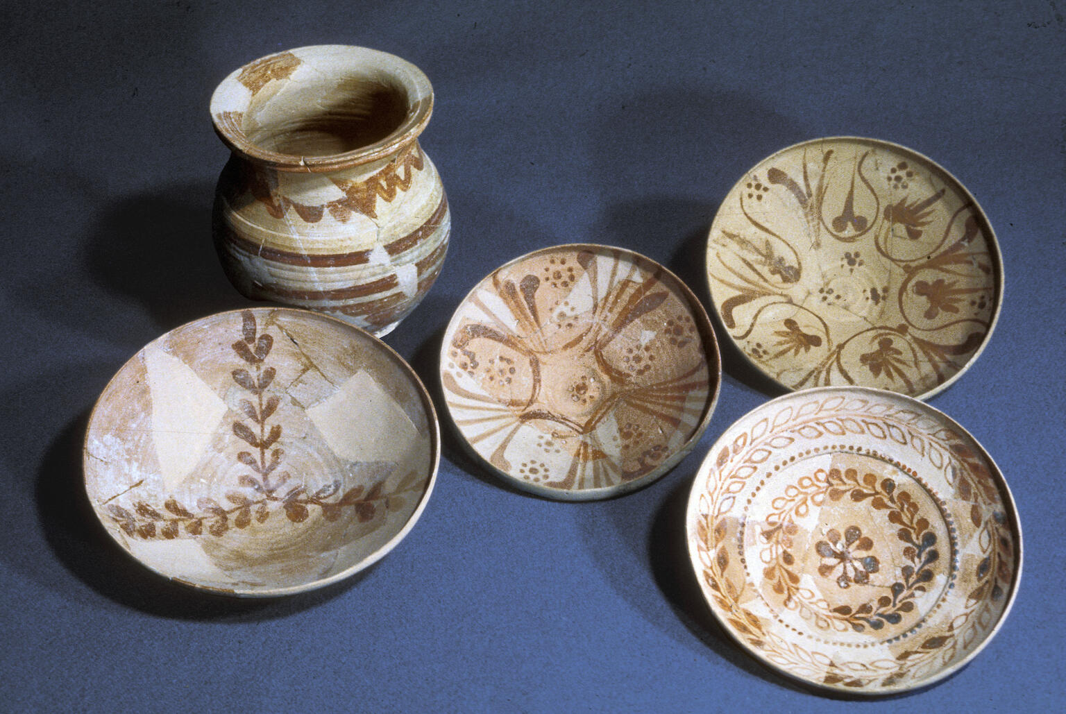 Five pieces of pottery are displayed on a flat surface: one small jar and four shallow bowls, all decorated with intricate brown geometric and floral patterns.