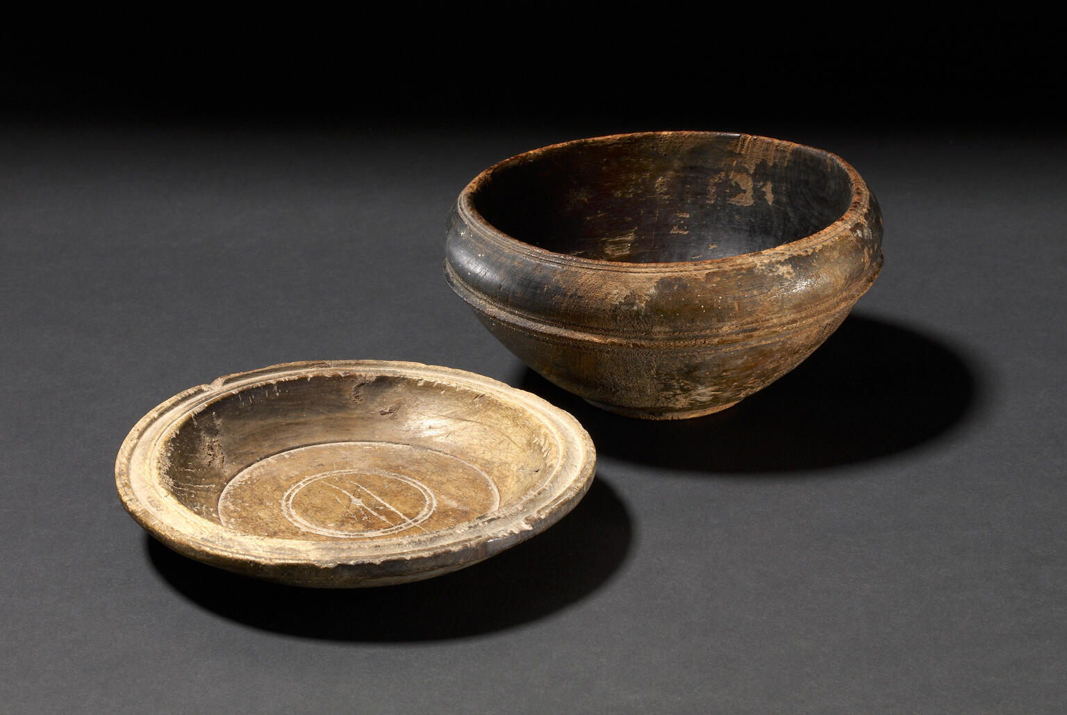 Two ancient wooden bowls on a dark background: one is deep and rounded with a blackish exterior, and the other is shallow and beige with a circular design etched inside.